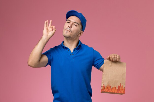 Вид спереди мужской курьер в синей форме держит пакет с едой на розовой стене, служба доставки униформы работника