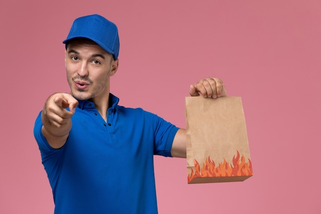 Front view male courier in blue uniform holding food package pointing out on pink wall, job worker uniform service delivery