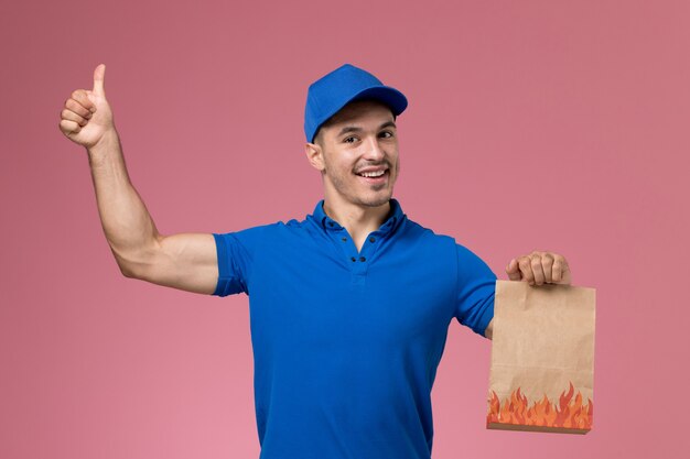 Курьер-мужчина в синей униформе, вид спереди, держит пакет с едой в виде знака на розовой стене, служба доставки униформы рабочего