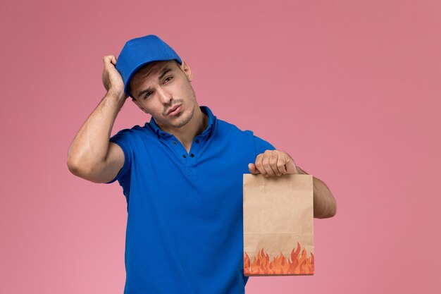 Front view male courier in blue uniform holding food paper package on pink wall, job worker uniform service delivery