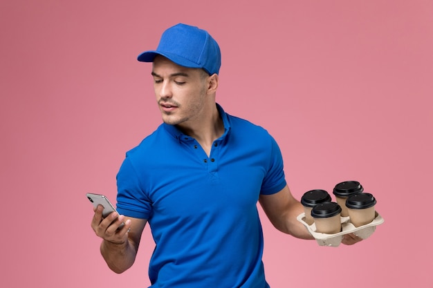 Front view male courier in blue uniform holding his phone and coffee cups on pink wall, uniform service delivery