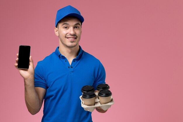 ピンクの壁に彼の電話のコーヒーカップを保持している青い制服の正面図男性宅配便、労働者の制服サービスの提供