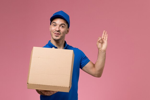 Front view male courier in blue uniform holding opening food delivery box on pink wall, uniform service delivery