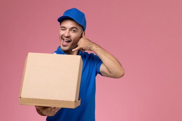 Front view male courier in blue uniform holding opening food delivery box on pink wall, uniform service delivery