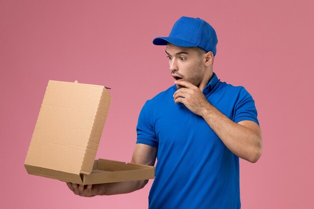 Front view male courier in blue uniform holding opening food delivery box on the pink wall, uniform service job delivery