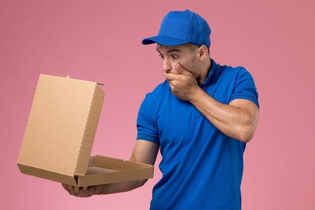 Front view male courier in blue uniform holding opening food delivery box with shocked expression on pink wall, uniform service job delivery
