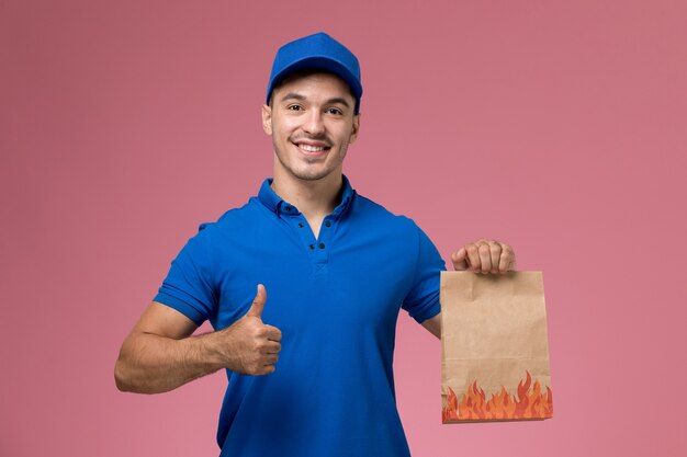 분홍색 벽, 균일 한 서비스 작업 배달에 미소로 종이 음식 패키지를 들고 파란색 제복을 입은 전면보기 남성 택배