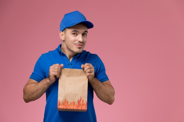 Front view male courier in blue uniform holding paper package on the pink wall, job worker uniform service delivery