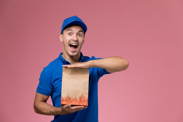 Front view male courier in blue uniform holding paper package rejoicing on pink wall, worker uniform service delivery