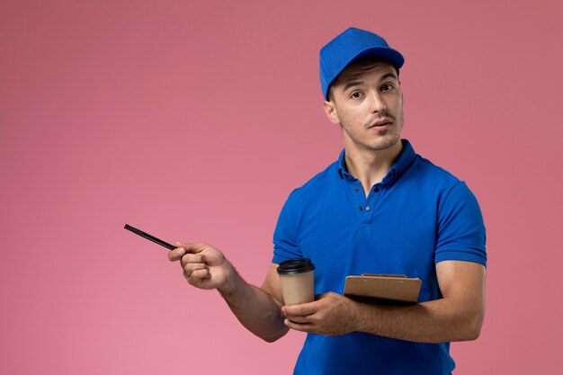 Вид спереди курьер-мужчина в синей форме, держащий ручку кофе вместе с блокнотом на розовой стене, служба доставки униформы работника