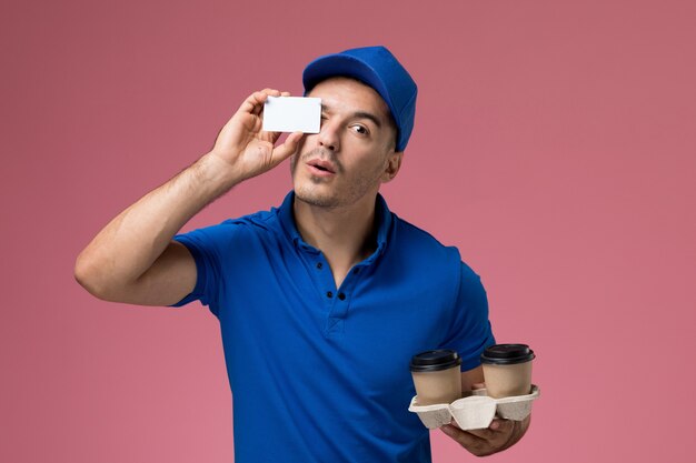 Front view male courier in blue uniform holding white card and coffee cups on pink wall, job worker uniform service delivery