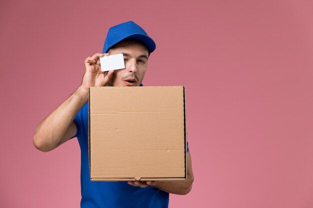 Вид спереди мужской курьер в синей форме, держащий белую карточную коробку с едой на розовой стене, служба доставки униформы работника