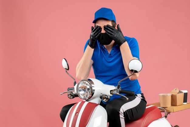 Front view male courier in blue uniform and mask closing his eyes on pink virus bike job delivery fast-food covid service work