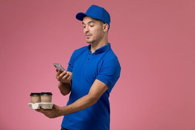 Front view male courier in blue uniform taking a photo of coffee on pink wall, worker uniform service delivery