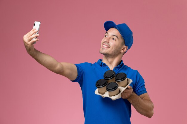 Курьер-мужчина, вид спереди в синей форме, делающий селфи с кофе на розовой стене, единообразная служба доставки на работу