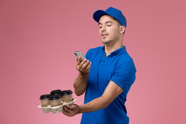 Front view male courier in blue uniform taking a shoot of coffee cups on pink wall, worker uniform service delivery