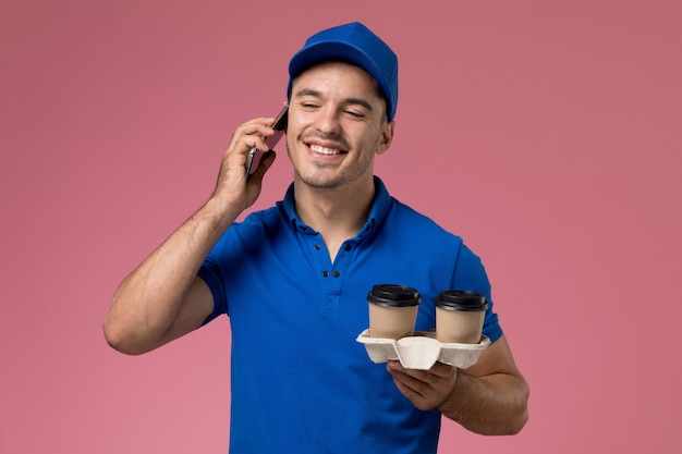 Corriere maschio di vista frontale in uniforme blu che parla sulle tazze di caffè del holdign del telefono sulla parete rosa, consegna di lavoro di servizio uniforme