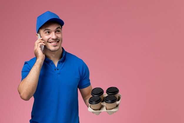 Front view male courier in blue uniform talking on the phone holding coffee on pink wall, uniform job worker service delivery