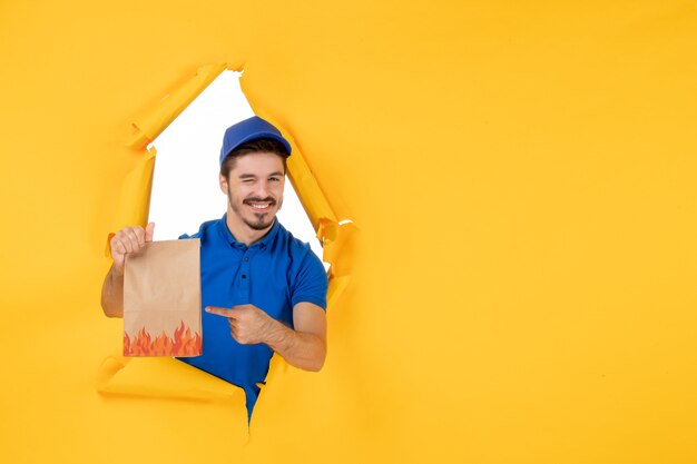 Вид спереди мужчина-курьер в синей форме с пакетом продуктов на желтом столе, служба доставки, работа, фото, цветная работа
