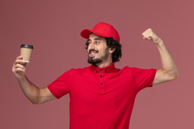 Front view male courier delivery man in red shirt and cape holding brown coffee cup and flexing on light-pink wall service uniform delivery employee