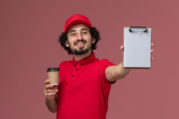 淡いピンクの壁に茶色のコーヒーカップとメモ帳を保持している赤いシャツとケープの正面図男性宅配便配達人サービス制服配達従業員の仕事