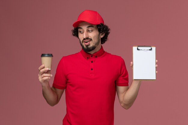 Front view male courier delivery man in red shirt and cape holding brown coffee cup and notepad on the light-pink wall service uniform delivery employee job