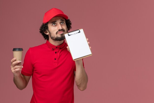 Front view male courier delivery man in red shirt and cape holding brown coffee cup and notepad thinking on the light-pink wall service uniform delivery employee