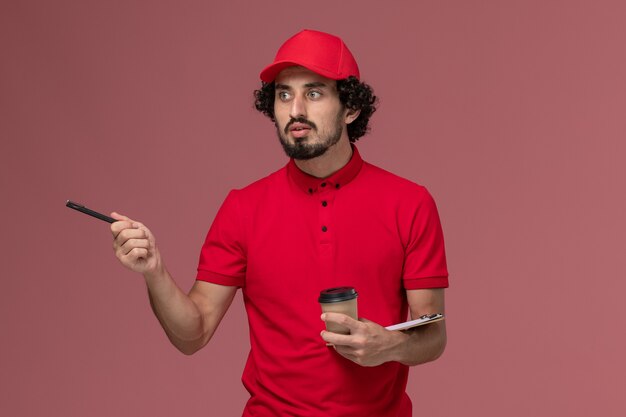 明るいピンクの壁のサービス配達従業員の仕事に茶色のコーヒーカップとペンでメモ帳を保持している赤いシャツとケープの正面図男性宅配便配達人