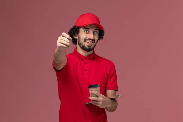 Front view male courier delivery man in red shirt and cape holding brown coffee cup and notepad with pen on light pink wall service delivery employee worker