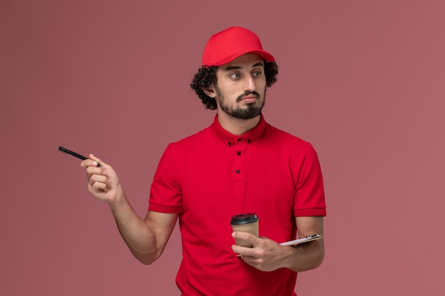 Uomo di consegna corriere maschio vista frontale in camicia rossa e mantello che tiene tazza di caffè marrone e blocco note con la penna sul dipendente di consegna servizio parete rosa chiaro