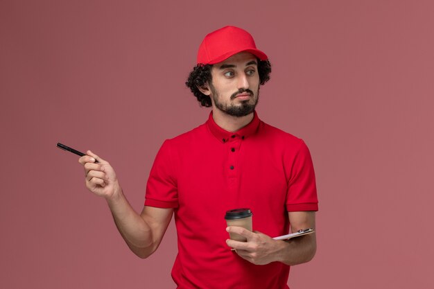 明るいピンクの壁のサービス配達従業員にペンで茶色のコーヒーカップとメモ帳を保持している赤いシャツとケープの正面図男性宅配便配達人