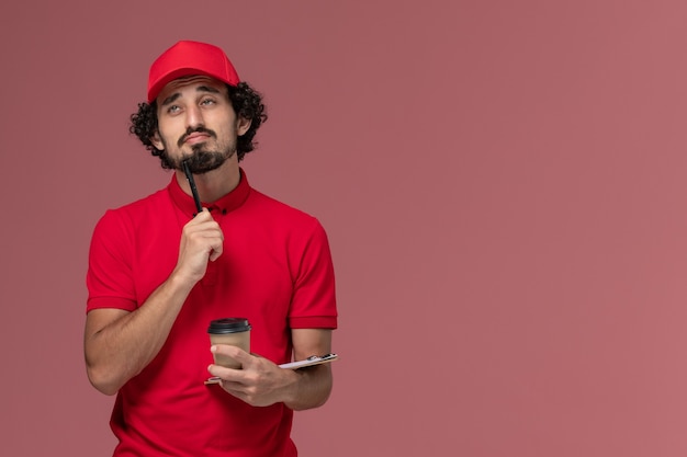 Вид спереди мужчина курьерской доставки в красной рубашке и накидке держит коричневую кофейную чашку и блокнот с ручкой, думая на светло-розовой стене, работа сотрудника службы доставки