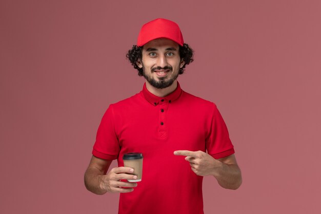 Вид спереди мужчина-курьер-доставщик в красной рубашке и плаще, держащий коричневую кофейную чашку на розовой стене, служба доставки униформа, сотрудник, работа, мужская работа