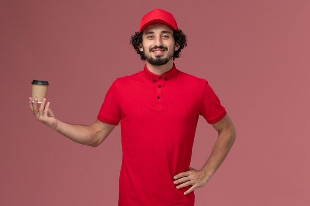 Uomo di consegna maschio del corriere di vista frontale in camicia rossa e mantello che tiene la tazza di caffè marrone sul lavoro maschio dell'operaio di lavoro dell'impiegato di consegna uniforme di servizio della parete rosa