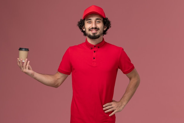 ピンクの壁に茶色のコーヒーカップを保持している赤いシャツとケープの正面図男性宅配便配達人サービス制服配達従業員ジョブワーカー男性の仕事