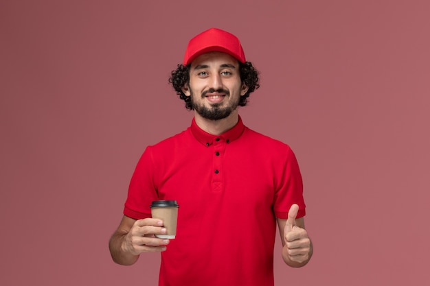 Uomo di consegna maschio del corriere di vista frontale in camicia rossa e mantello che tiene la tazza di caffè marrone sul lavoro dell'operaio di lavoro dell'impiegato di consegna uniforme di servizio della parete rosa