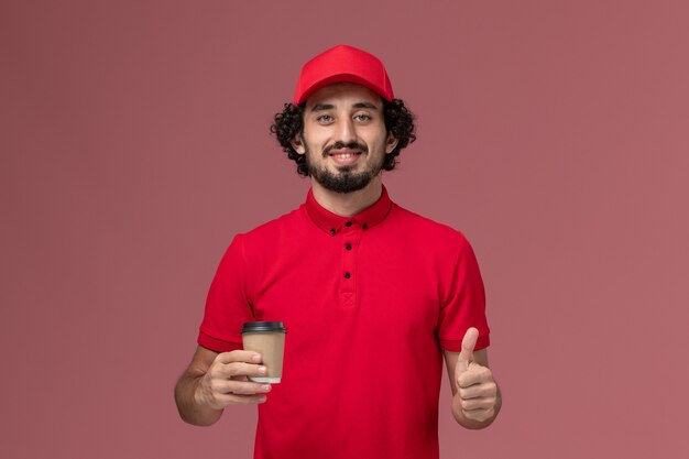 Front view male courier delivery man in red shirt and cape holding brown coffee cup on pink wall service uniform delivery employee job worker work