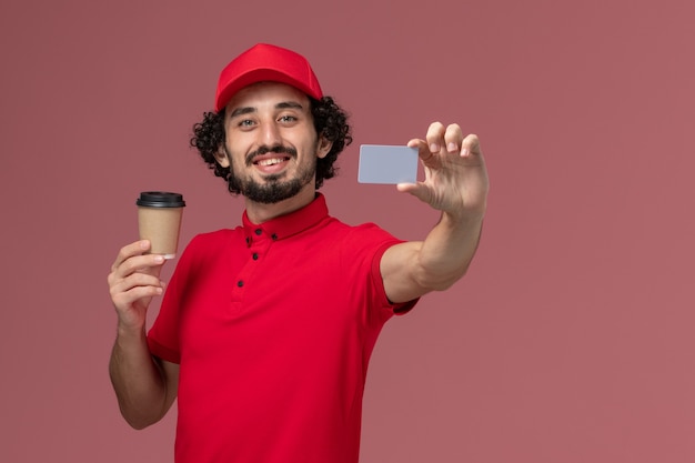 Front view male courier delivery man in red shirt and cape holding brown coffee cup with grey card on light-pink wall service uniform delivery employee male