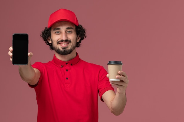Вид спереди мужчина-курьер-доставщик в красной рубашке и плаще, держащий коричневую кофейную чашку доставки и телефон на светло-розовой стене служащий службы доставки