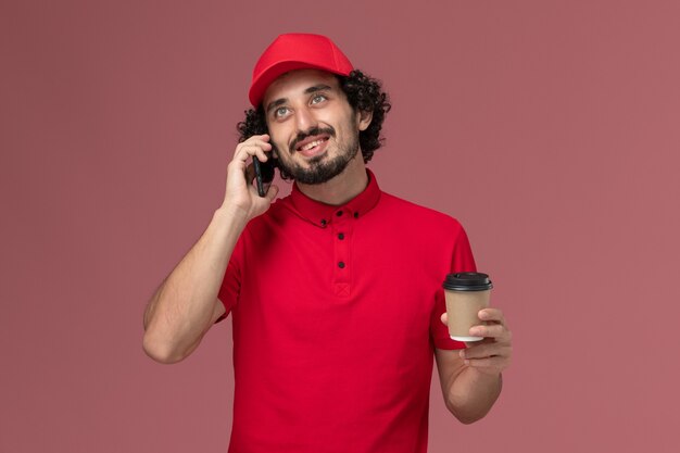Front view male courier delivery man in red shirt and cape holding brown delivery coffee cup and talking on the phone on light pink wall service delivery employee work