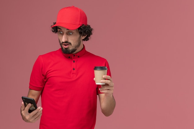 Uomo di consegna corriere maschio vista frontale in camicia rossa e mantello che tiene tazza di caffè consegna marrone e utilizzando un telefono sul dipendente di consegna servizio parete rosa chiaro