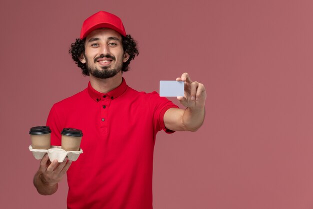 Вид спереди мужчина курьерской доставки в красной рубашке и накидке с коричневыми кофейными чашками и серой карточкой на светло-розовой стене сотрудника службы доставки