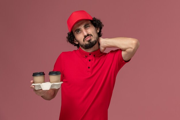 밝은 분홍색 벽 서비스 배달 직원에 목이 아픈 갈색 배달 커피 컵을 들고 빨간 셔츠와 케이프에 전면보기 남성 택배 배달 남자