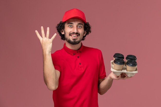 Front view male courier delivery man in red shirt and cape holding brown delivery coffee cups on the light pink wall job service delivery employee work