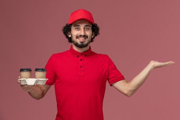 Вид спереди мужчина курьерской доставки мужчина в красной рубашке и плаще держит коричневые кофейные чашки доставки на светло-розовой стене сотрудник службы доставки мужская работа