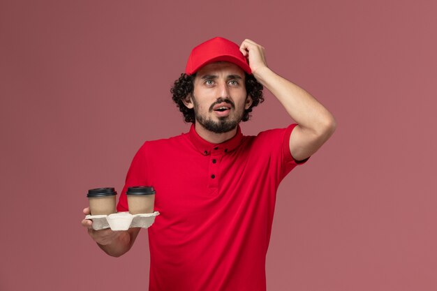 淡いピンクの壁に茶色の配達コーヒーカップを保持している赤いシャツとケープの正面図男性宅配便配達人男性サービス配達従業員男性