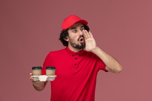 Front view male courier delivery man in red shirt and cape holding brown delivery coffee cups on light-pink wall service delivery employee work worker