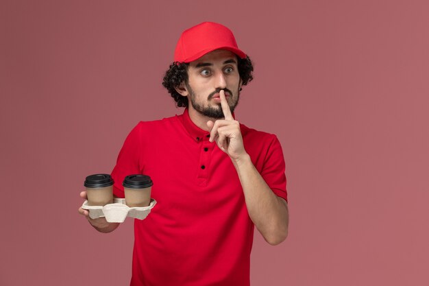 Вид спереди мужчина курьерской службы доставки в красной рубашке и накидке, держащий коричневые кофейные чашки на светло-розовой стене, работа сотрудника службы доставки