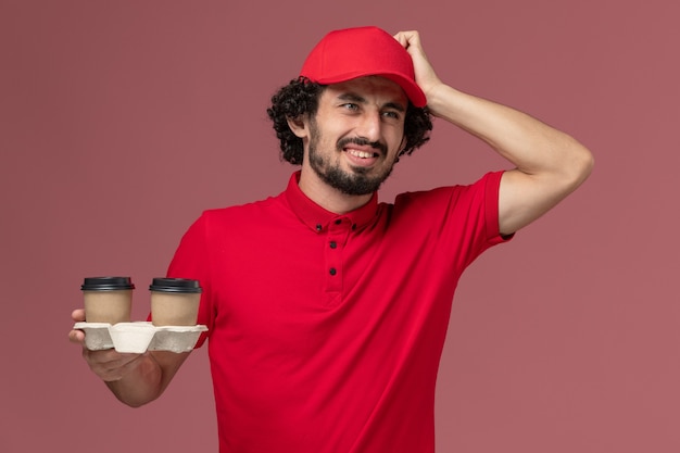 Front view male courier delivery man in red shirt and cape holding brown delivery coffee cups on the light-pink wall service delivery employee