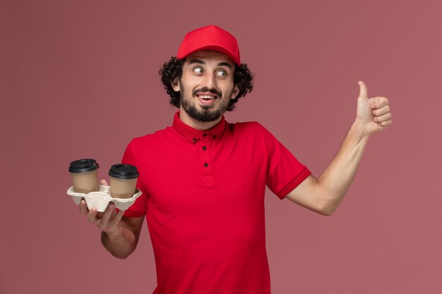 Вид спереди мужчина-курьер-курьер в красной рубашке и плаще, держащий коричневые кофейные чашки на светло-розовой стене, служащий службы доставки